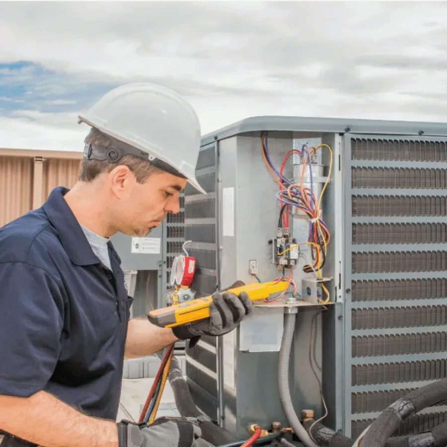 HVAC technician in a hard hat testing an outdoor air conditioner with a multimeter